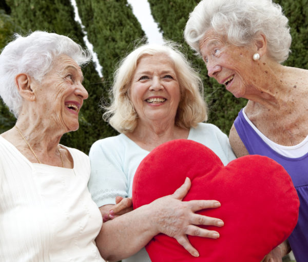 three senior woman standing together; middle one holds a red heart pillow